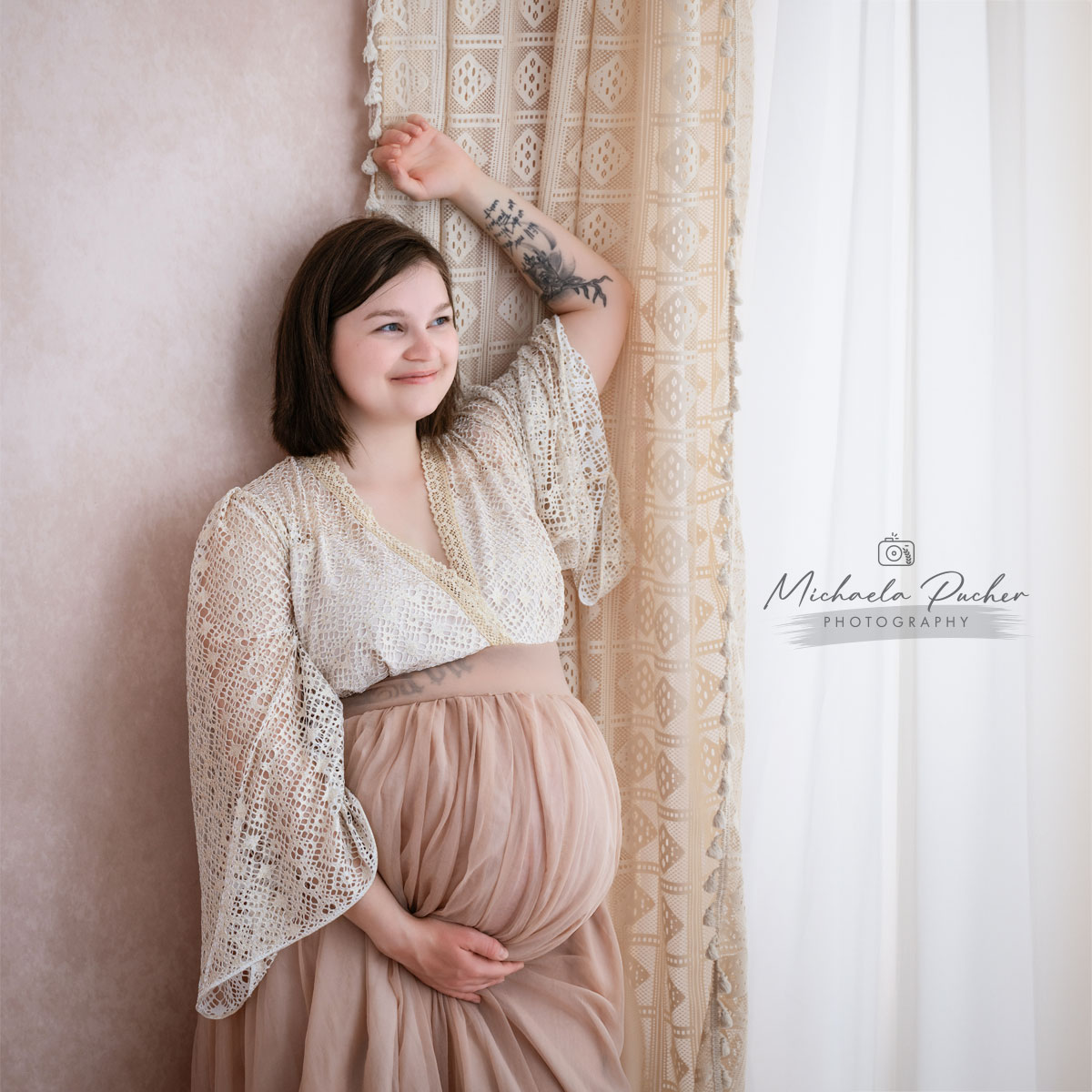 Schwangere Frau lehnt lächelnd an einem cremefarbenen Vorhang, hält ihren Babybauch und blickt sanft zur Seite.